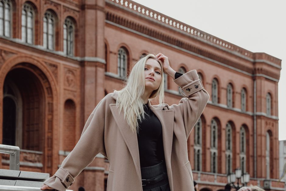 Fashionable woman with blonde hair poses stylishly in front of a historic brick building.