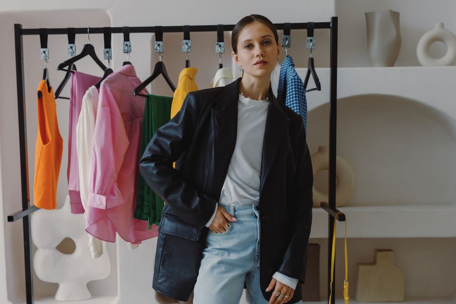 Stylish woman in a black leather jacket poses beside a clothing rack in a chic boutique.