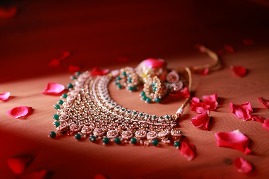 Close-up of a gold necklace with green stones, surrounded by red rose petals on a wooden surface.