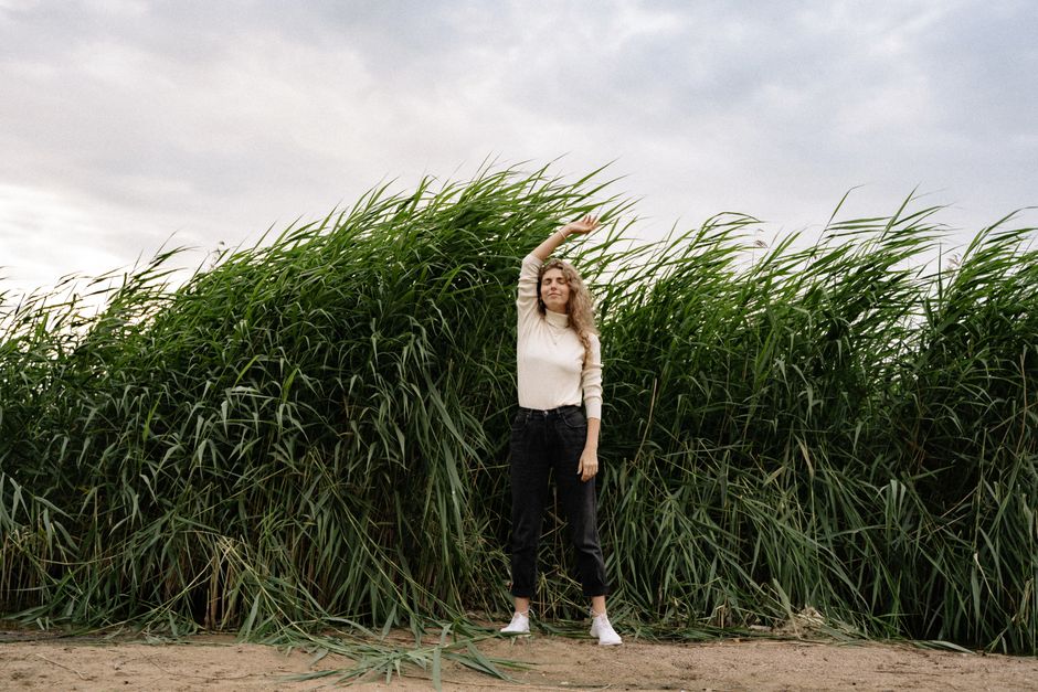 A woman with long hair poses with arm raised in front of tall grass and clouds.