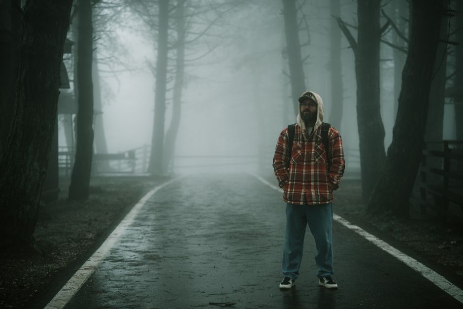 A solitary man in a plaid jacket stands on a foggy forest path, creating a moody atmosphere.