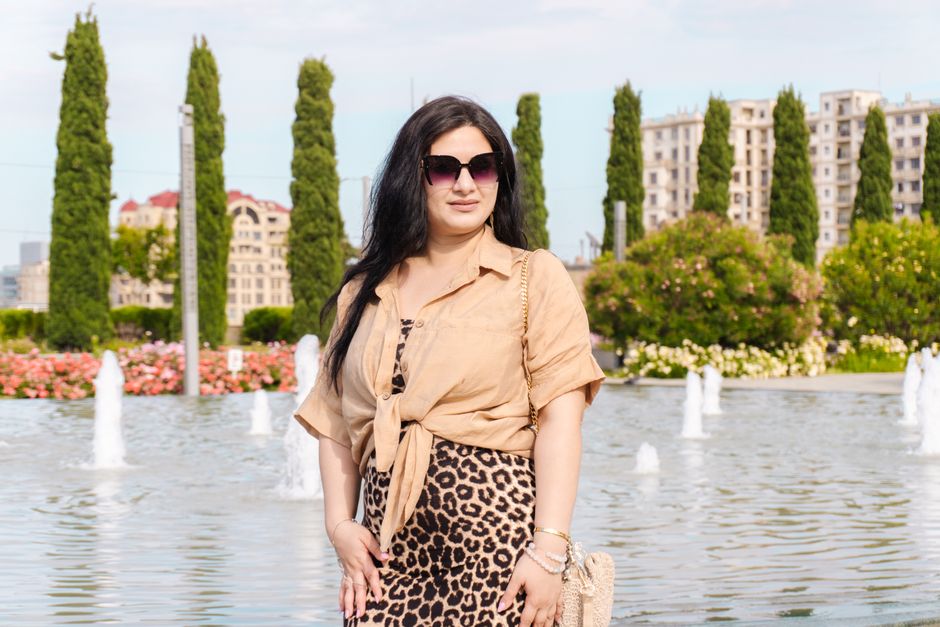 Stylish woman in sunglasses posing by a fountain in a city park with tall trees and buildings in the background.