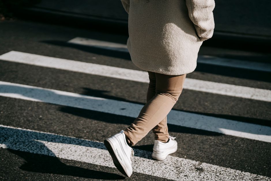 Back view crop unrecognizable female wearing casual outerwear crossing road on pedestrian crosswalk on sunny day