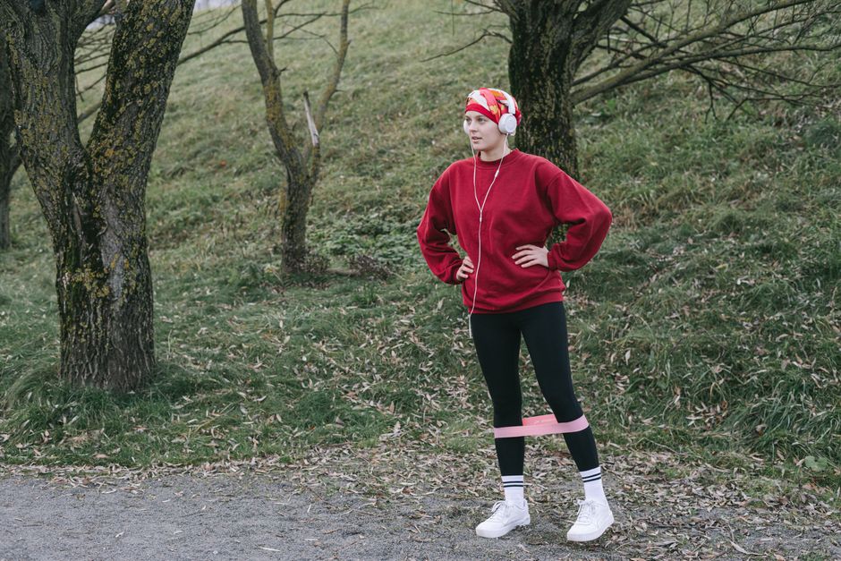 Woman in red sweatshirt exercising with resistance band outdoors, wearing headphones.