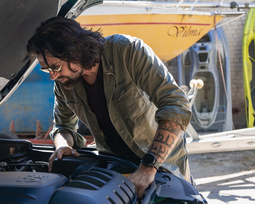 Bearded man checking car engine in sunny outdoor space with boats in background.