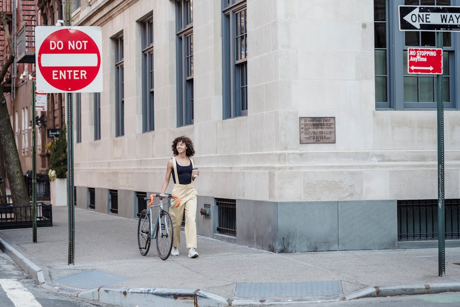 Smiling ethnic female with hot drink to go and bicycle strolling on urban walkway between restrictive signs while looking away