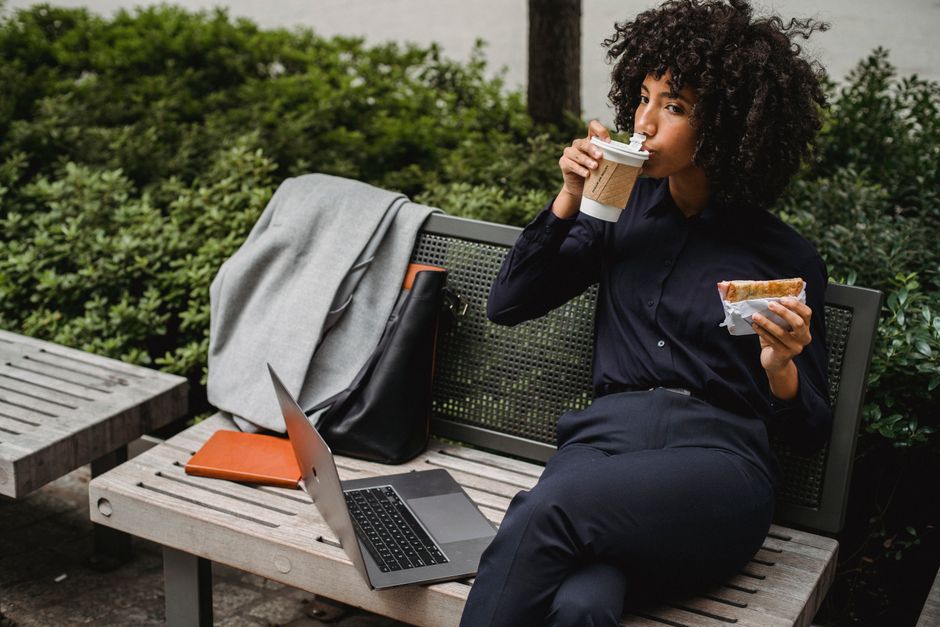Adult woman sipping coffee and eating a sandwich while working outdoors on a bench.