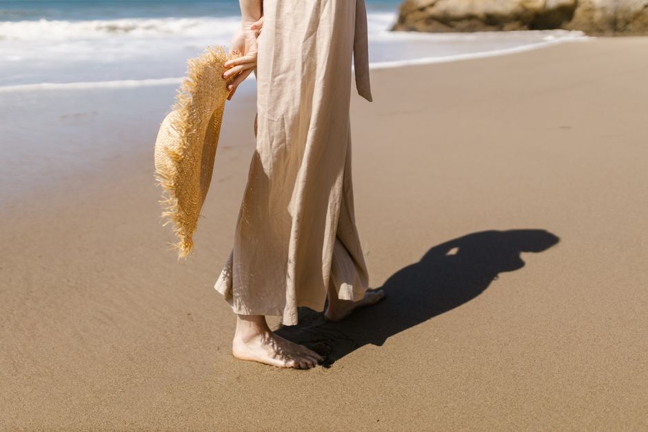 A woman walks barefoot on a sandy beach holding a straw hat, casting a shadow.