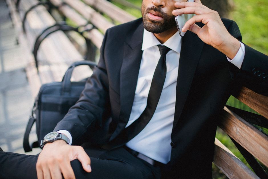 A bearded businessman in a black suit seated on a bench, using a smartphone outdoors.