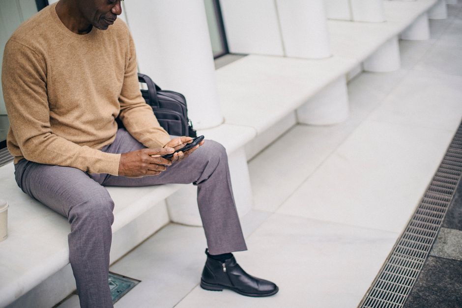 Crop black mature man sitting on stone bench in downtown and text messaging on smartphone