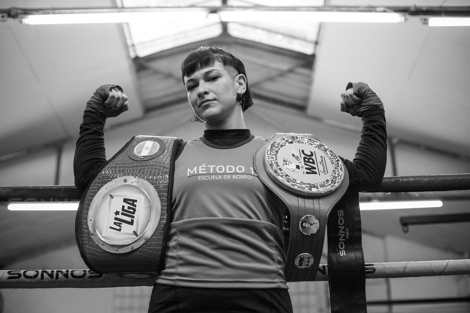 A female boxer proudly showcases her championship belts in a boxing gym.