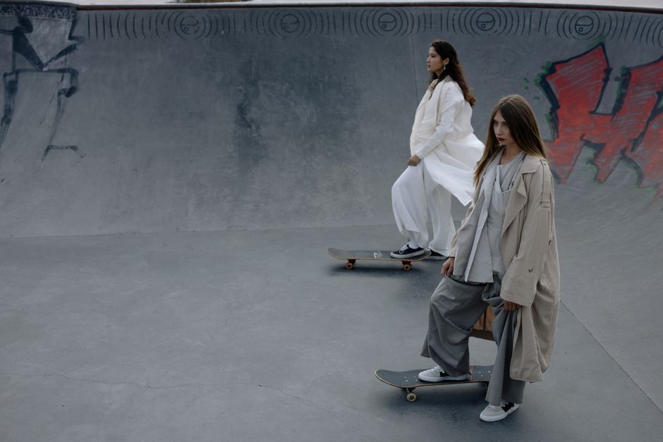 Two women in fashionable clothing skateboarding in an urban concrete skatepark.