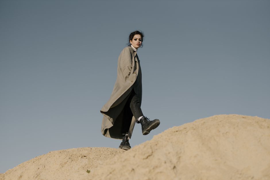 A fashionable woman in a trench coat walks across a sandy terrain under a clear sky.