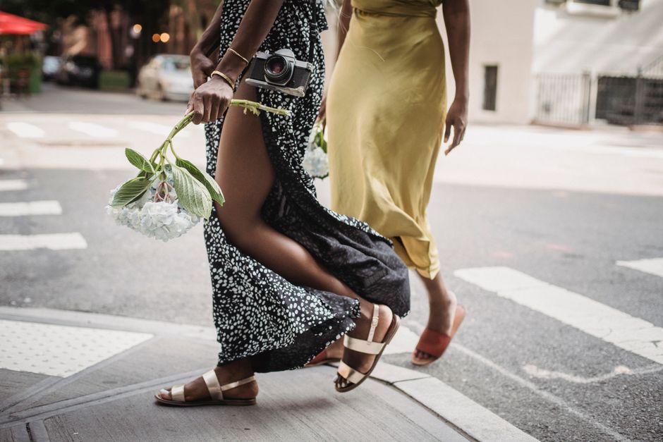 Two women in elegant dresses walk stylishly across an urban street.