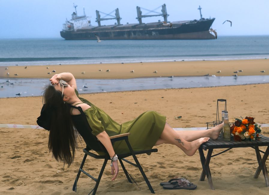 A woman in a green dress relaxes on a beach chair with a distant cargo ship, conveying leisure and maritime allure.