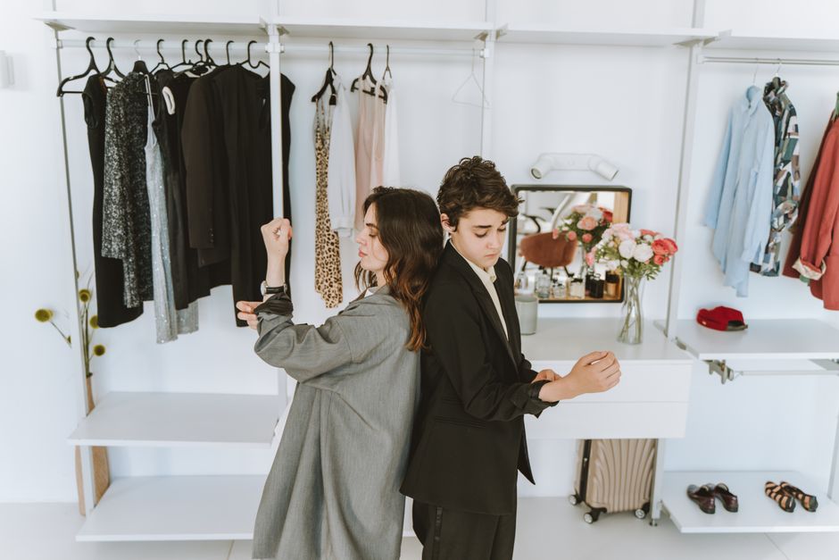 A mother and son dressed in black and gray suits, stand back to back in a stylish wardrobe setting.
