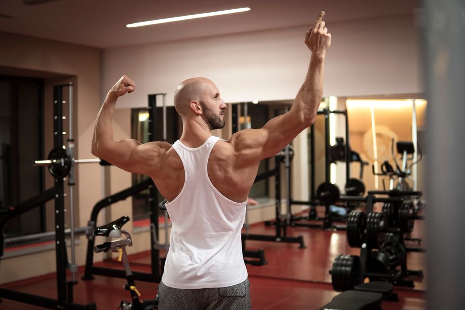 A muscular man in a gym flexes his back muscles wearing a white tank top.