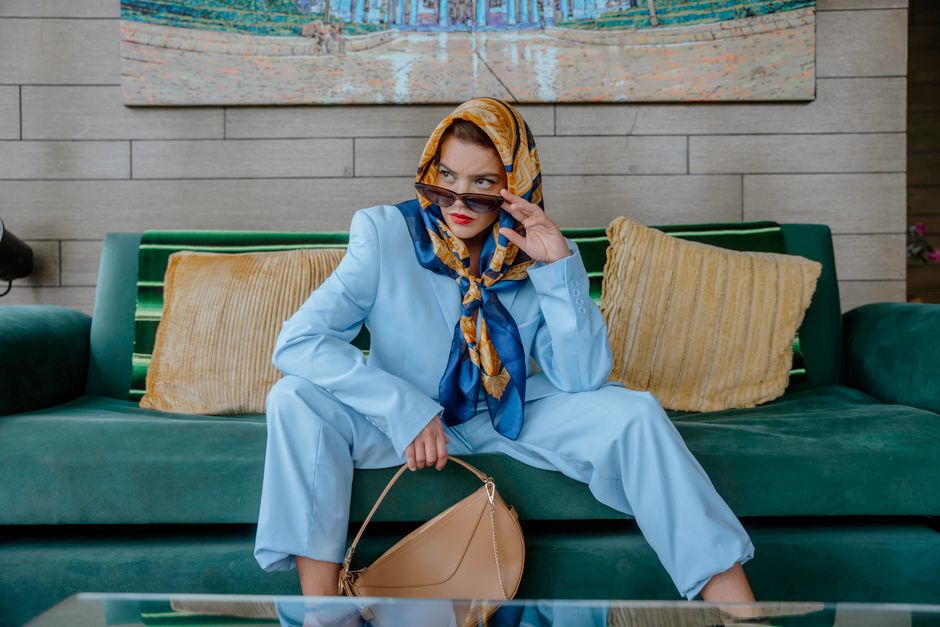 Elegant woman in a blue suit and scarf sitting stylishly on a sofa inside a living room.