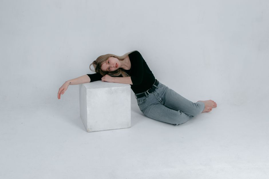 A woman in jeans leans against a white cube in a minimalist studio setting, barefoot and relaxed.
