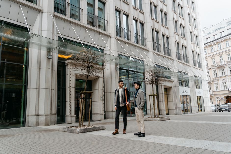 Two businessmen outdoors in conversation near modern office buildings, wearing formal attire.