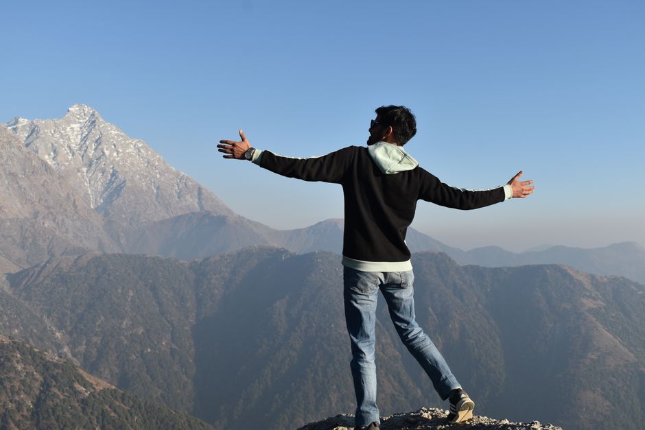 A man stands embracing nature, overlooking majestic mountains under a clear blue sky.