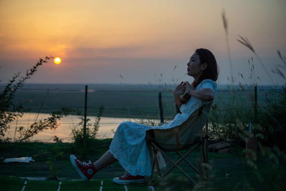 A woman relaxes in a chair outdoors, gazing at a beautiful sunset near a river.