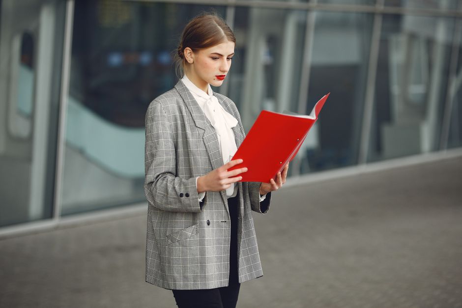 Young female entrepreneur in stylish formal wear reading documents while standing near modern business building in city street