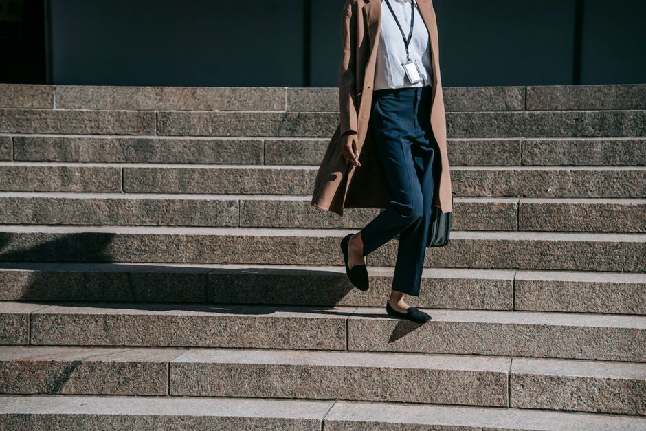 Crop anonymous female entrepreneur in brown coat with badge and handbag walking down stairs