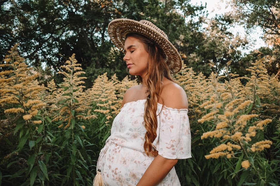 Pregnant woman in a floral dress with a straw hat in a sunny field, eyes closed peacefully.