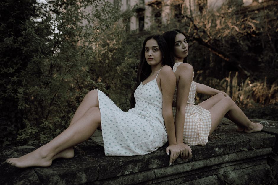 Artistic outdoor portrait of two women in dresses sitting on a stone wall surrounded by greenery.