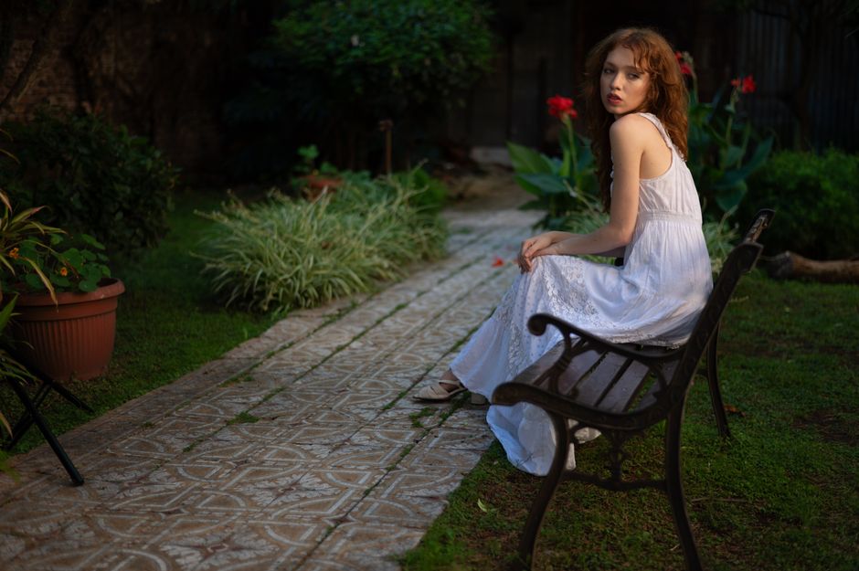 A woman in a white dress sits on a garden bench during twilight surrounded by lush greenery.