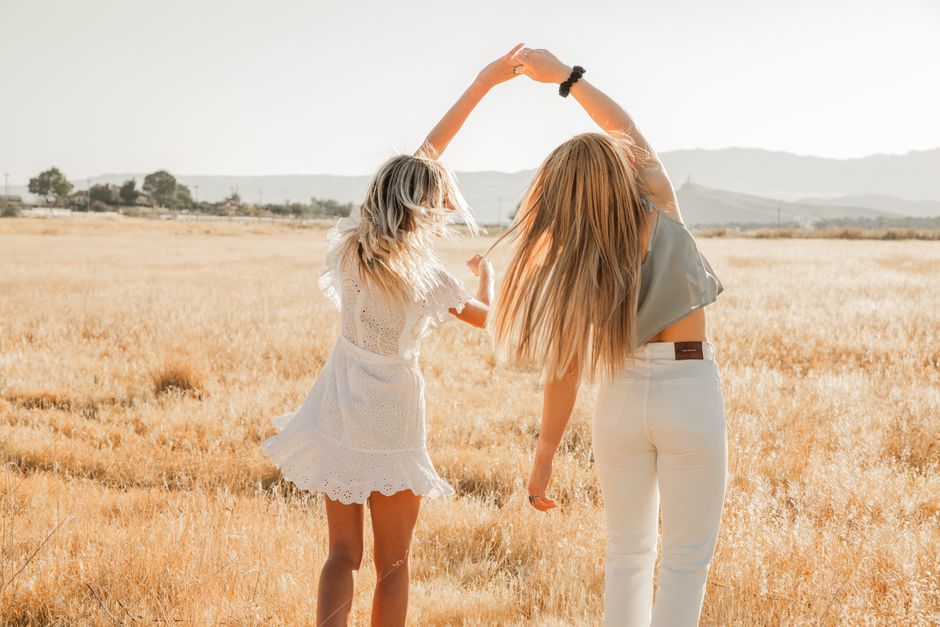 Back view of anonymous women in stylish outfit standing with raised arms on grass meadow behind mounts in back lit