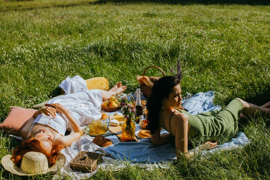 Two women enjoying a relaxing summer picnic on a grass field with fresh food and drinks.