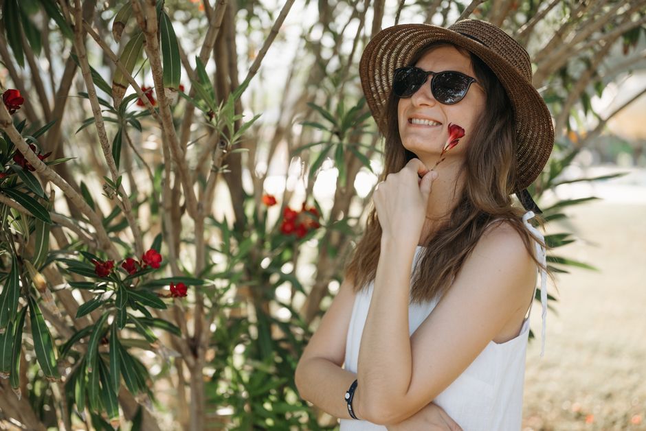 A woman in a sun hat smiling beside blooming red flowers in a garden.
