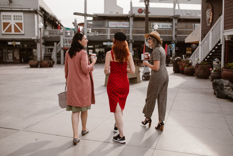 Back view of stylish middle aged female friends in elegant outfits drinking takeaway coffee and chatting while walking on city street during trip