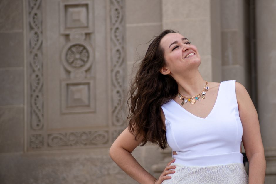A smiling young woman in a white dress stands joyfully against an ornate stone wall.