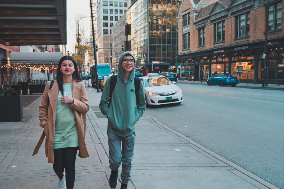 Two young adults walking on a city street surrounded by buildings and traffic.