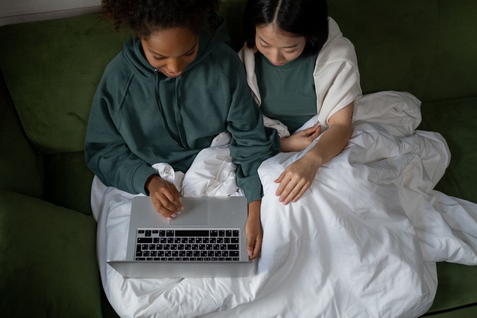 A couple relaxing on a sofa under a white blanket, using a laptop in a cozy setting.
