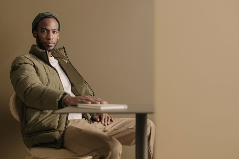 Stylish man in olive jacket sitting indoors with minimalistic background.