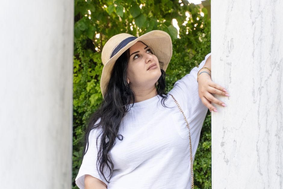 Woman in a light outfit with a straw hat poses by a column in a lush garden setting.