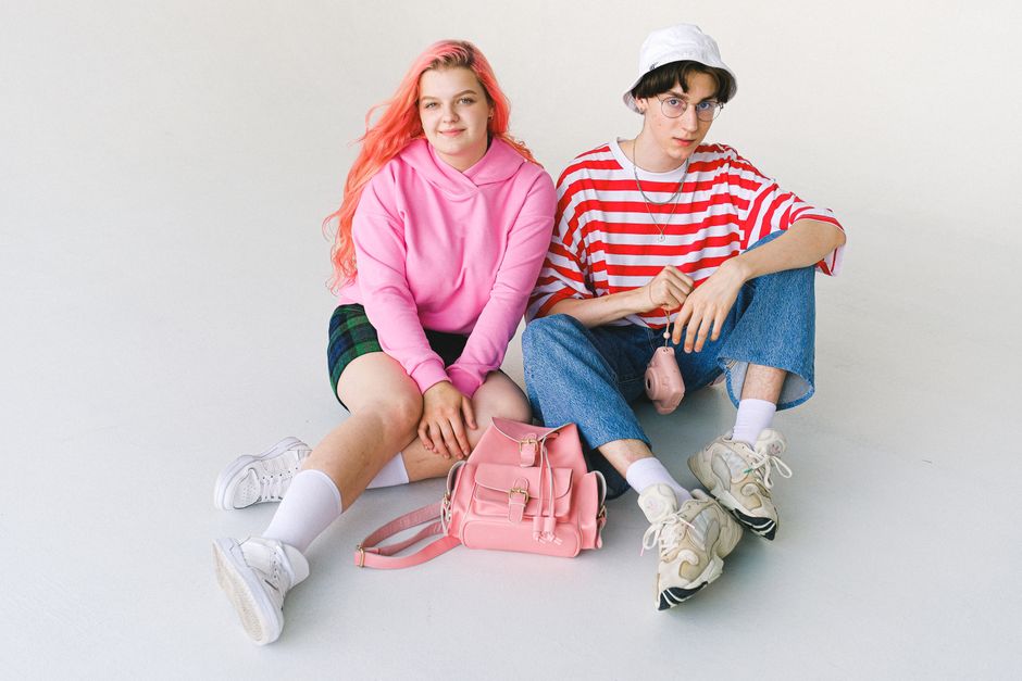 From above of full body informal teenager couple wearing colorful clothes sitting on floor with camera and backpack against white background and looking at camera