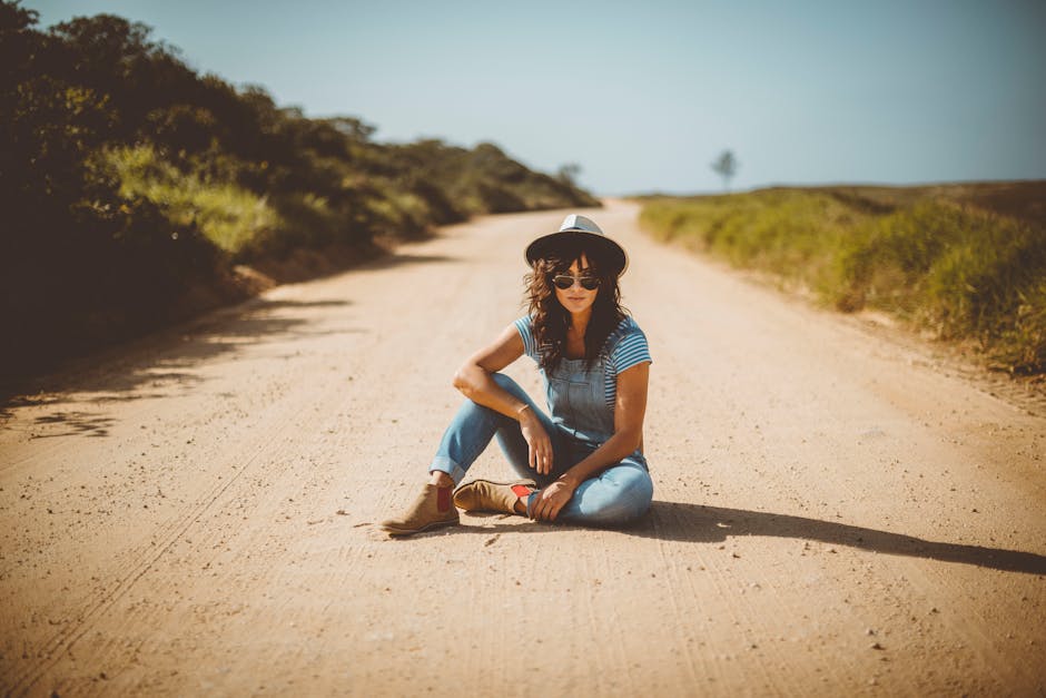 Casual young woman wearing sunglasses sitting on a dirt road enjoying a sunny day.