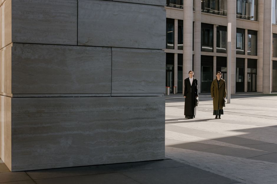 Two women in formal attire walk along a modern city street with a prominent building backdrop.